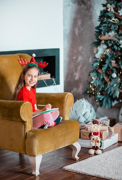 Une jolie fille dans un chapeau de Père Noël écrit une lettre au Père Noël près de l'arbre de Noël Enfance heureuse