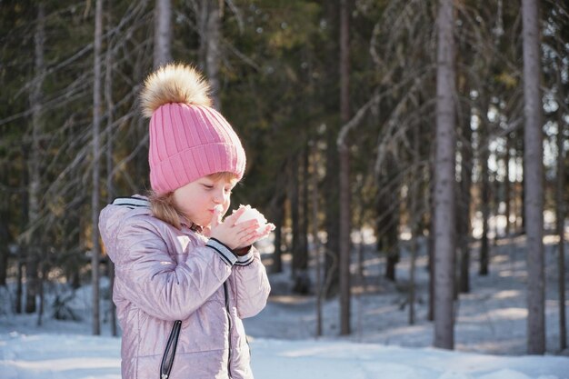 Jolie fille dans un chapeau et une veste s'amuse avec des flocons de neige par temps ensoleillé d'hiver dans ses paumes