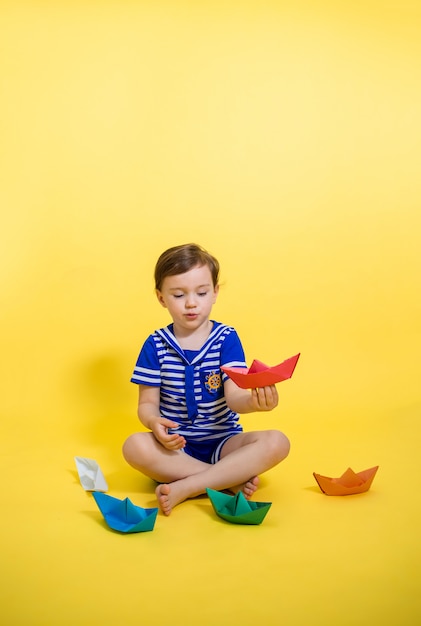 Une jolie fille dans un costume de marin joue avec des bateaux en papier sur un espace jaune. Orientation verticale.