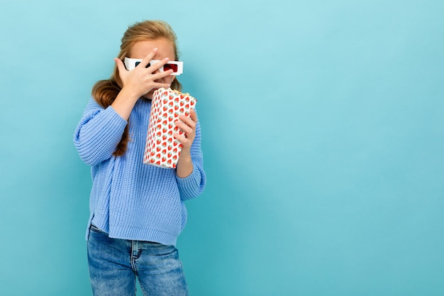 Jolie fille dans des lunettes de cinéma tient du pop-corn dans ses mains sur bleu clair