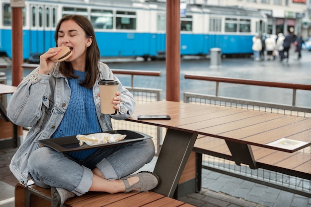 Jolie fille dans un style décontracté mange un hamburger avec du café assis sur la terrasse d'été d'un café