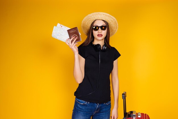 Une jolie fille dans un t-shirt noir et des lunettes part en voyage avec une grande valise détient un passeport et des billets