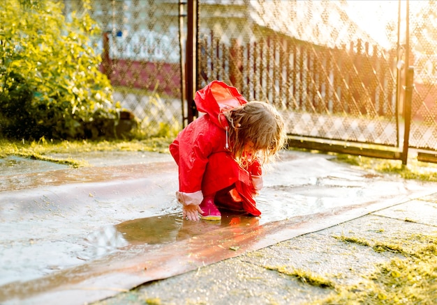 Jolie fille dans une veste rouge saute dans la flaque d'eau.Le cadre chaud d'été ou soleil d'automne. été dans le village.