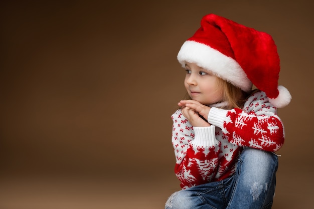 Jolie fille dans des vêtements confortables et un chapeau de Noël se trouve sur le sol, photo isolée sur fond marron