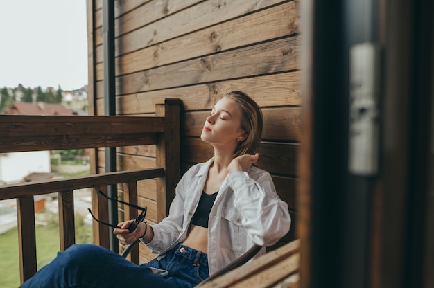 Jolie fille dans des vêtements décontractés est assise sur le balcon dans une chaise et s'amuse les yeux fermés