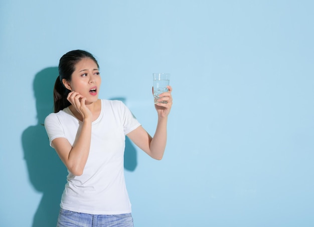 jolie fille douce regardant l'eau glacée s'inquiéter de l'attaque des dents sensibles et faire des émotions douloureuses debout sur fond de mur bleu.