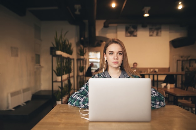 Jolie fille avec des écouteurs et des vêtements décontractés travaille sur un ordinateur portable dans le café