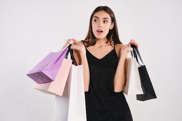 Jolie fille élégante choquée en robe noire tenant des sacs à provisions étonnamment en détournant les yeux sur fond blanc isolé