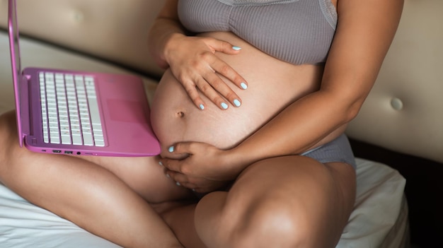 Jolie fille enceinte parcourt l'ordinateur portable et touche doucement son ventre. Gros plan femme mains sur ordinateur portable avec gros ventre grossesse avancée.