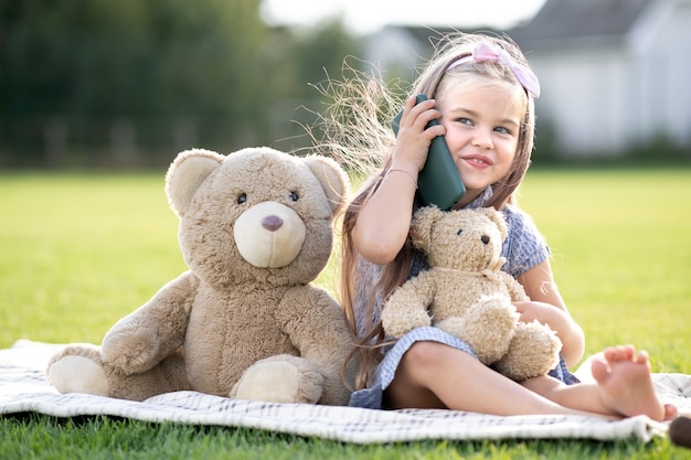 Jolie fille enfant assise dans un parc d'été sur une pelouse d'herbe verte avec son ours en peluche parlant au téléphone portable à l'extérieur en été.