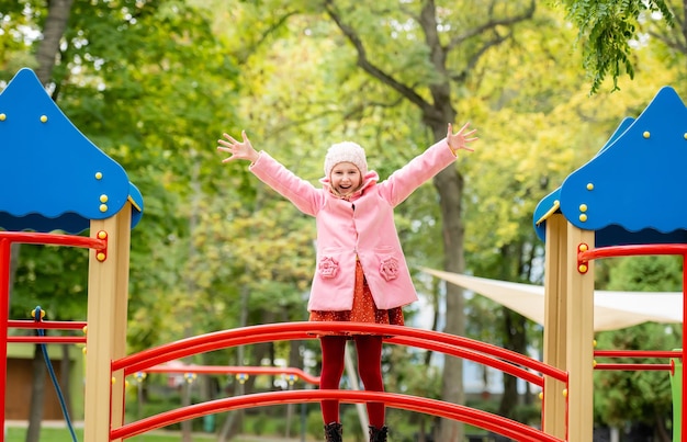 Jolie fille enfant debout sur une aire de jeux avec les mains au jour d'automne à l'extérieur et souriante enfant de sexe féminin...