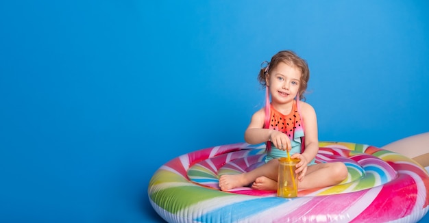 Jolie fille enfant en maillot de bain buvant du jus d'orange assis sur un matelas gonflable coloré sur bl...