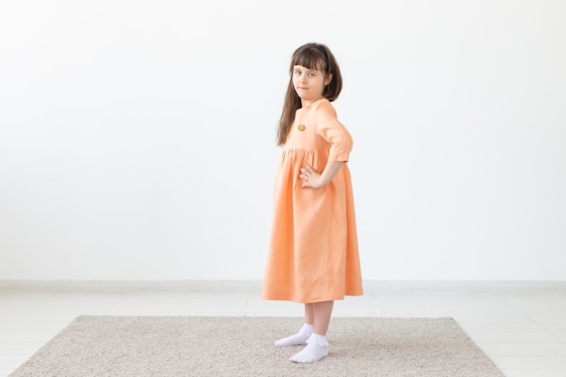 Jolie fille enfant posant pour la caméra, dans une robe courte orange, mur blanc avec espace copie