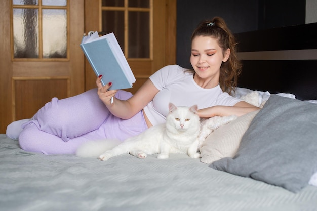 Une jolie fille est allongée sur le lit avec un livre et caresse un chat blanc