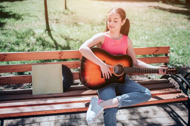 Jolie Fille Est Assise Sur Un Banc Et Joue De La Guitare. Elle Regarde Vers Le Bas Le Vynil Et Sa Couverture Qui Est également Sur Le Banc. Elle Profite Du Moment.