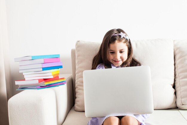 La jolie fille a eu une idée. La petite fille assise sur un canapé avec un ordinateur portable et des livres