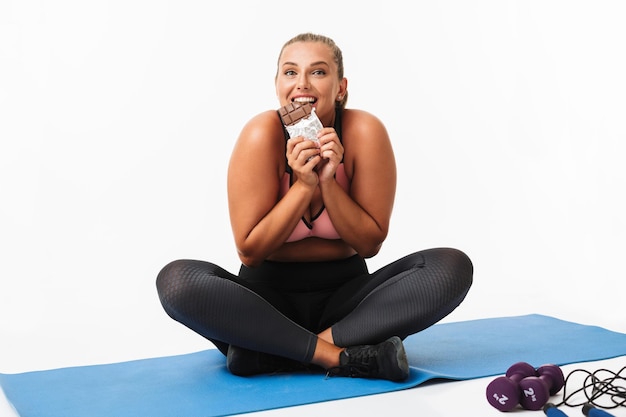 Jolie fille avec un excès de poids en haut sportif et leggings assis sur un tapis de yoga mangeant joyeusement du chocolat regardant à huis clos sur fond blanc isolé