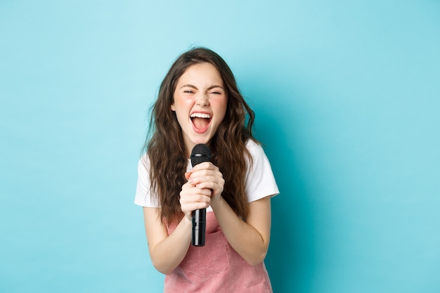 Jolie fille excitée chantant un karaoké, tenant un microphone et souriant heureux, debout sur fond bleu.
