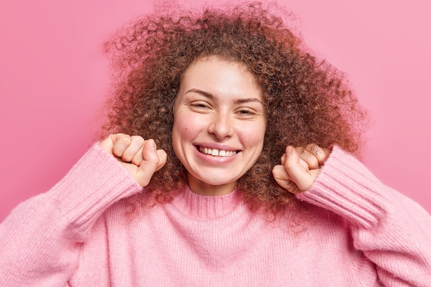 Une jolie fille féminine aux cheveux bouclés et touffus serre les poings, sourit avec joie, a l'air heureuse et détendue, porte un pull décontracté isolé sur un mur rose. Concept d'émotions et de sentiments sincères des gens