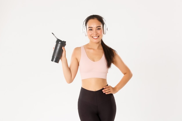 Jolie fille de fitness asiatique mince et en forme dans les écouteurs, écouter de la musique pendant l'entraînement, boire de l'eau ou des protéines de bouteille, fond blanc.