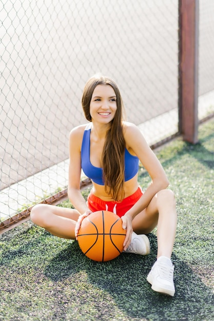 Photo une jolie fille en forme est assise avec un ballon de basket sur le terrain de sport.