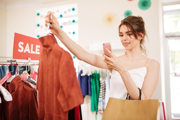 Jolie fille en haut blanc debout près du porte-vêtements de vente avec un T-shirt à la main et en prenant une photo sur son téléphone portable dans la boutique. Portrait d'une femme souriante avec des sacs à provisions à la main dans un magasin de vêtements