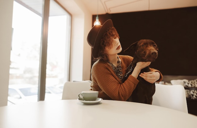 Jolie fille hipster dans des vêtements à la mode est assise à une table dans un café avec un chien dans ses bras
