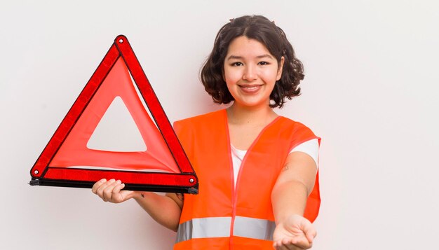 Jolie fille hispanique souriant joyeusement avec amical et offrant et montrant un triangle d'accident de voiture concept