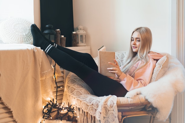 Jolie fille imaginant le monde extraordinaire du livre en position couchée dans un fauteuil rond confortable près d'une fenêtre à l'intérieur