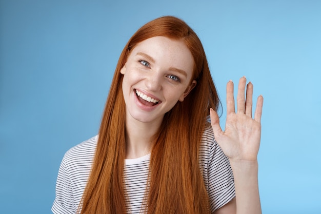 Jolie fille impertinente rousse confiante pure peau propre yeux bleus inclinant la tête gaiement en agitant la main bonjour salut geste saluant vous regardez ami accueillant sympathique caméra, fond de studio debout