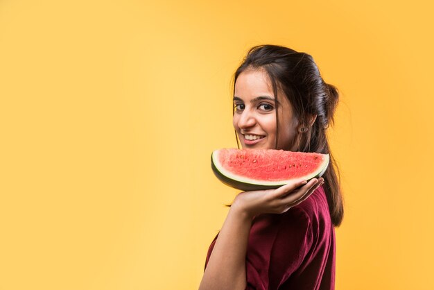 Jolie fille indienne ou asiatique tenant une tranche de pastèque ou de Tarbuja en hindi, debout isolée sur fond blanc ou jaune.