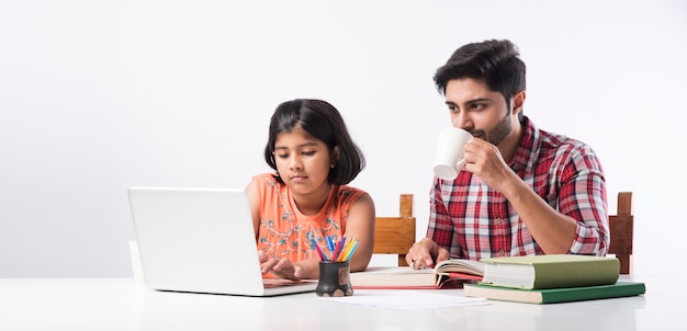 Jolie fille indienne avec son père étudiant ou faisant ses devoirs à la maison à l'aide d'un ordinateur portable et de livres - concept d'enseignement en ligne