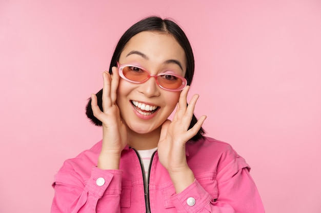 Jolie fille japonaise moderne à lunettes de soleil souriante et heureuse posant sur fond rose dans des vêtements élégants