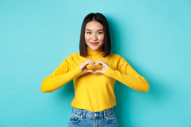 Jolie fille japonaise en pull jaune, montrant le geste du cœur et disant je t'aime, regardant sincèrement la caméra, debout sur fond bleu