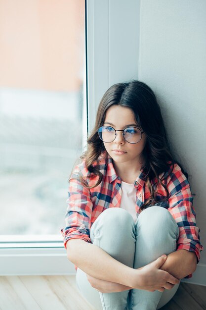 Jolie fille en jeans et chemise assise près de la fenêtre avec ses bras autour des genoux et regardant au loin