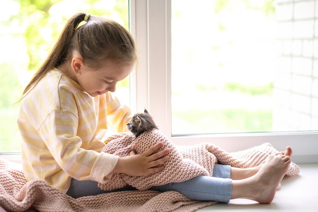 Une jolie fille joue avec son petit chaton moelleux assis sur le rebord de la fenêtre de la maison Amitié avec un animal de compagnie