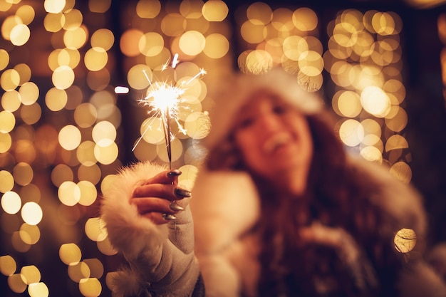 Jolie fille joyeuse avec des cierges magiques s'amusant à la veille de Noël dans la rue de la ville avec beaucoup de lumières de vacances.