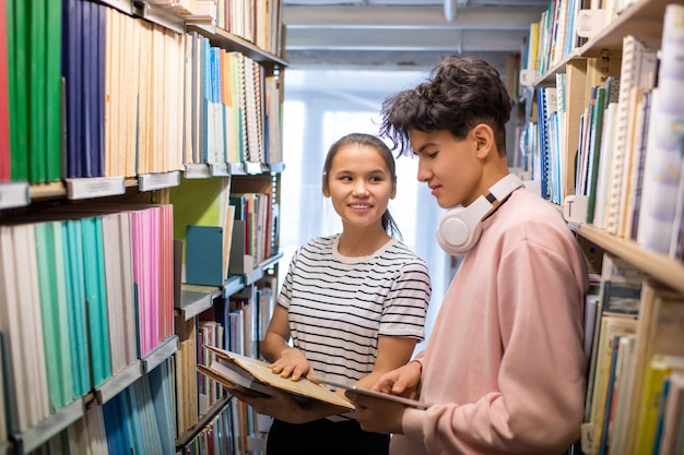 Jolie fille avec des livres en regardant son camarade de classe avec tablette au cours de la discussion de l'affectation à domicile entre les étagères de la bibliothèque