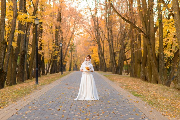 Jolie fille en longue robe de mariée blanche posant dans un chemin rural parmi les arbres d'automne en forêt dans l'atmosphère de l'heure d'or.