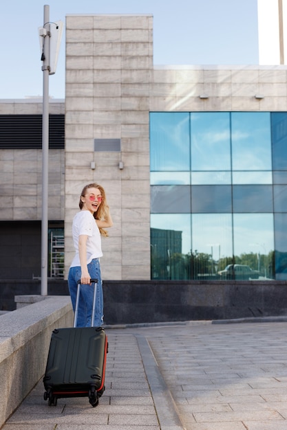 Jolie fille avec des lunettes avec des bagages va sur la route en été