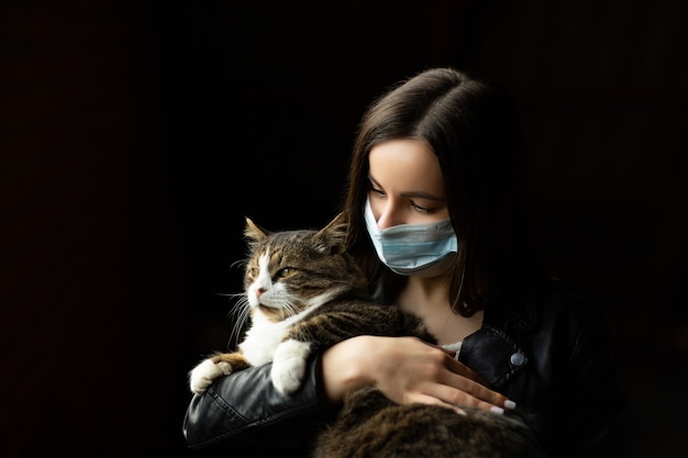 Jolie fille en masque médical tenant un chat sur un mur noir