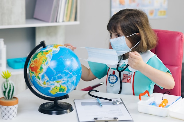 Une jolie fille médecin met un masque sur le globe de la planète Terre Une petite fille vêtue d'un costume de médecin traite la planète Terre.