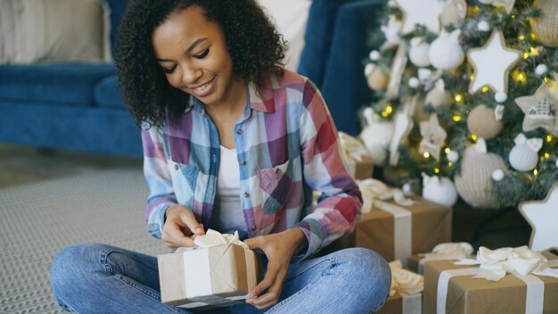 Photo jolie fille métisse emballant une boîte-cadeau près de l'arbre de noël à la maison