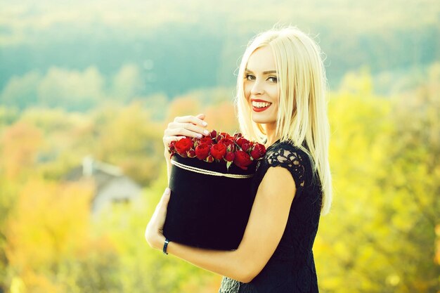 Jolie fille mignonne ou belle femme blonde en robe noire sexy avec des fleurs roses rouges dans une boîte en plein air sur fond naturel