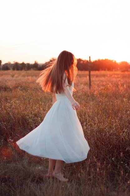 Jolie fille mignonne tourne dans une robe blanche sur le terrain. Rayons de soleil. Coucher de soleil orange. Concept. Une femme