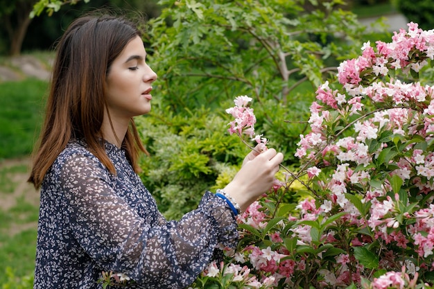 Jolie fille mince portant une robe élégante debout dans un parc fleuri se penchant touchant la fleur