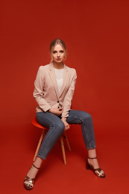 Jolie fille modèle glamour attrayant portant un blazer beige et un jean assis sur la chaise, isolé sur fond rouge