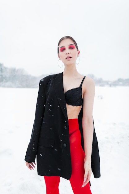 Jolie fille modèle de lingerie dans des vêtements à la mode avec un blazer et un soutien-gorge collants rouges posant à l'extérieur avec de la neige dans la nature