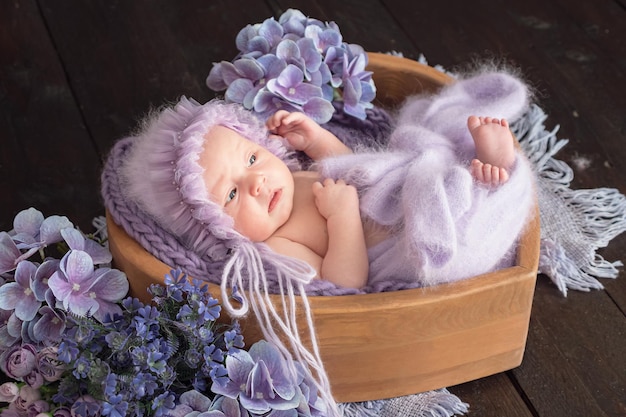 Jolie fille nouveau-née en bonnet de laine doux mord les doigts se trouvant dans le panier parmi les fleurs violettes sur fond sombre Séance photo bébé en studio gros plan extrême