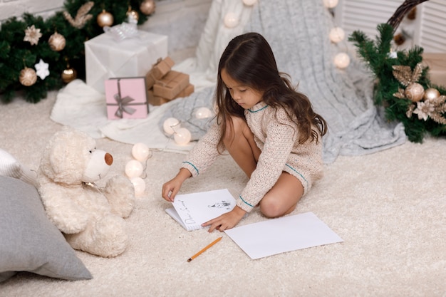 Jolie fille avec ours en peluche écrivant une lettre au père Noël à la maison